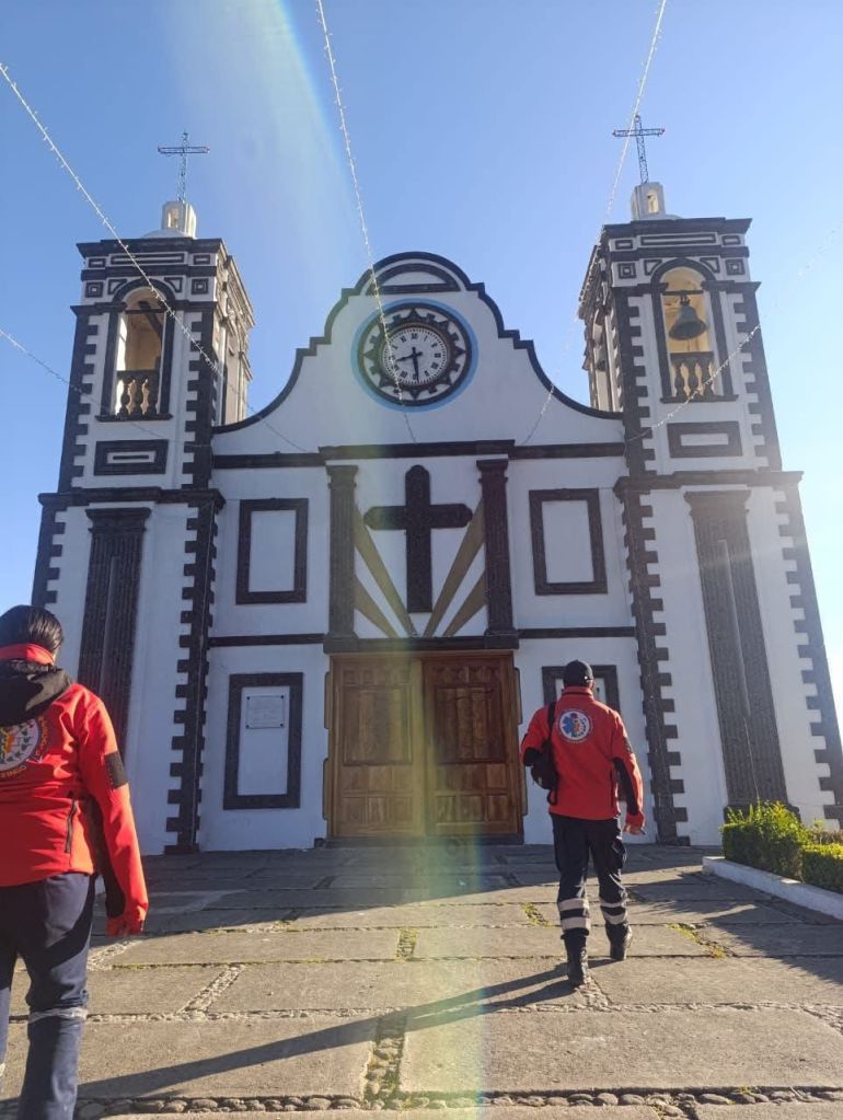 Vista de una iglesia con un reloj, donde dos personas vestidas con chaquetas rojas caminan hacia la entrada.