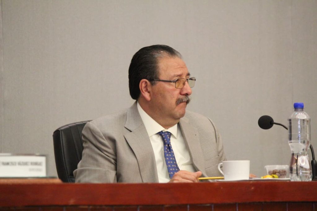 Hombre vestido con traje claro sentado en una mesa de reunión, mirando hacia un lado, con vaso de agua y taza frente a él.