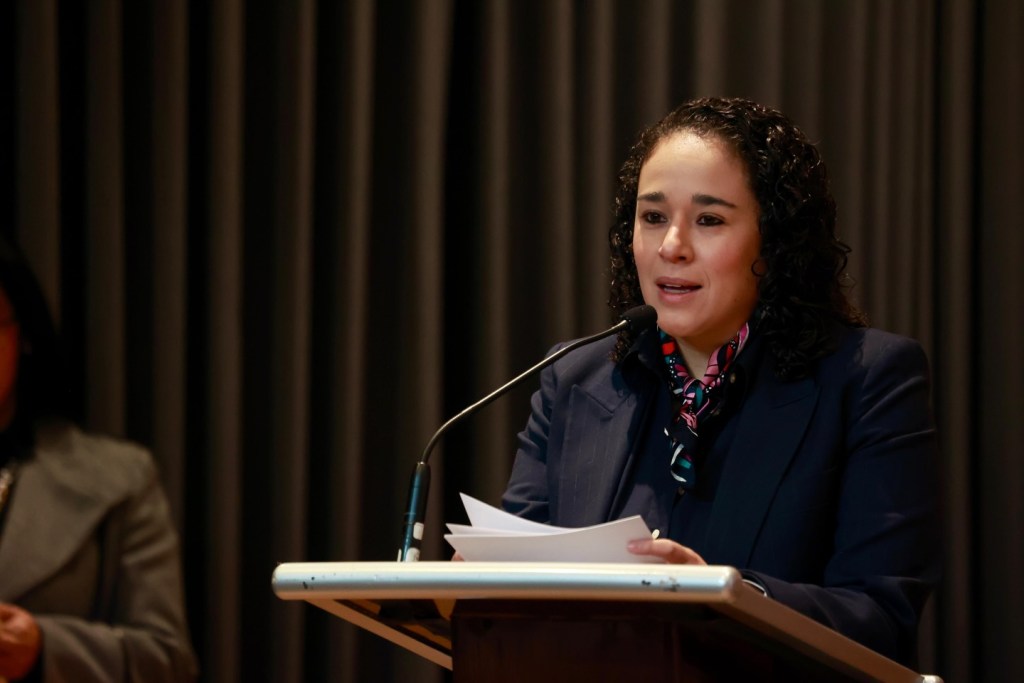 Una mujer hablándole a una audiencia desde un podio, sosteniendo papeles en la mano, con un fondo de cortinas de color oscuro.