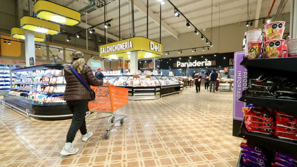 A shopper pushing an orange cart in a grocery store aisle, with various food items displayed in refrigerated sections. Signs indicate the deli and bakery areas.