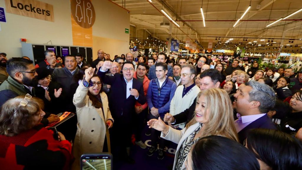 A large crowd of diverse individuals gathered indoors, some raising their hands in excitement, while others smile, in a lively atmosphere, possibly during a public event or gathering.