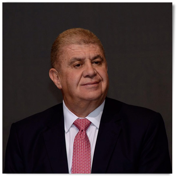 Retrato de un hombre con cabello corto y canoso, vestido con un traje oscuro y corbata roja, mostrando una expresión serena.