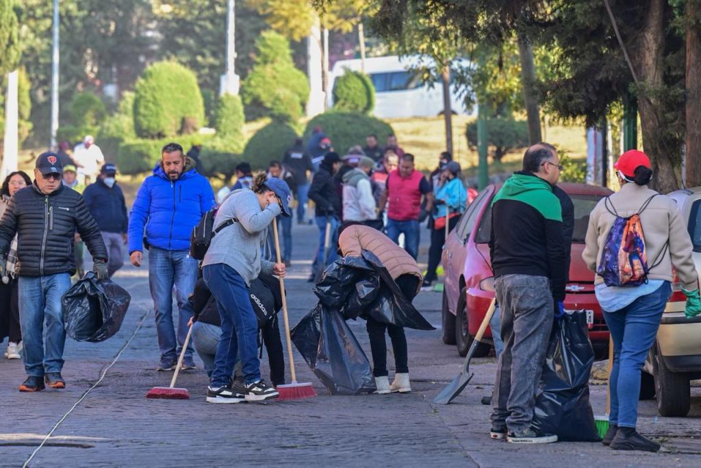 Un grupo de voluntarios participa en una jornada de limpieza, recogiendo basura y barriendo en una calle de Toluca, mientras otros se encuentran en el fondo.