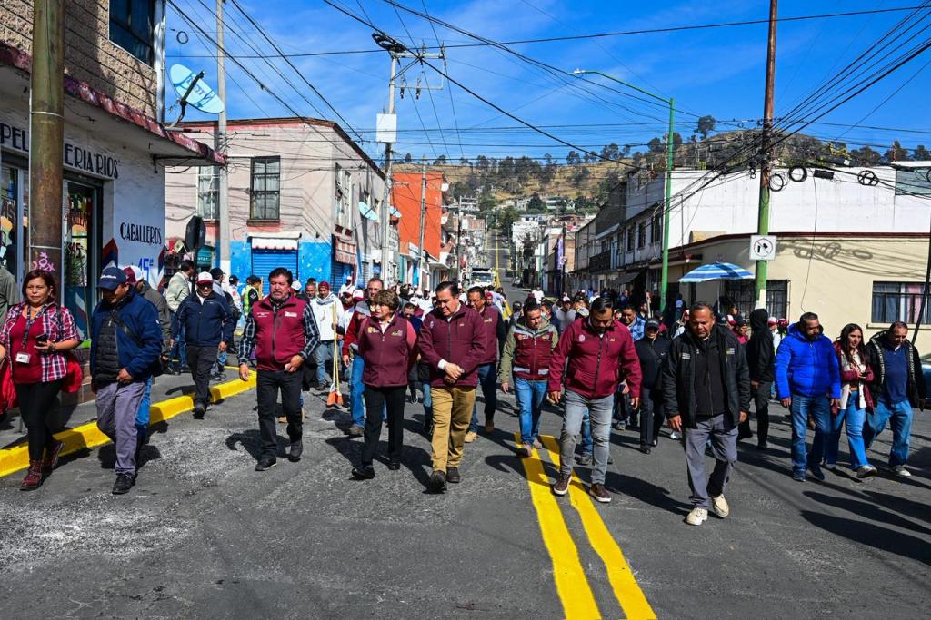 Una multitud de personas camina por una calle en Toluca, participando en la campaña de limpieza 'Limpiemos Nuestro EdoMéx', liderada por la gobernadora Delfina Gómez Álvarez, con edificios y una mañana soleada de fondo.
