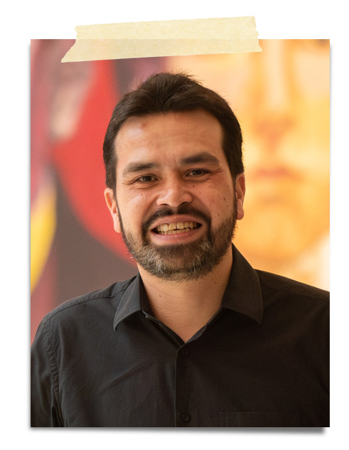 Retrato de un hombre sonriendo, con barba y cabello oscuro, vistiendo una camisa negra. Fondo desenfocado con colores cálidos.