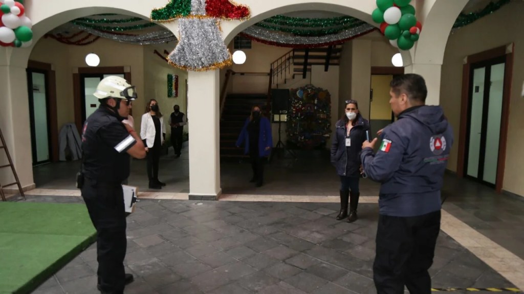 Grupo de personas en un edificio, donde dos hombres con casco y chaquetas realizan una revisión de seguridad, con decoración festiva visible en el techo.
