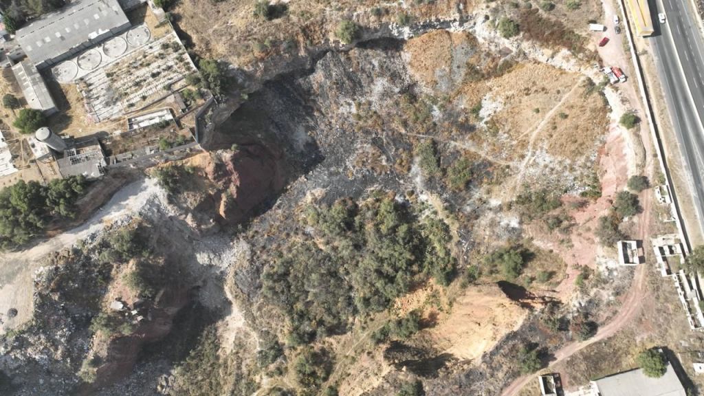 Aerial view of a barren landscape with a cliff, vegetation, and a nearby industrial building, showcasing a mix of dirt, rubble, and sparse greenery.