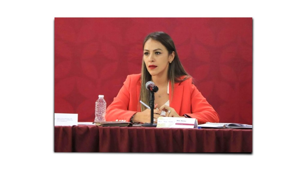 Mujer con chaqueta roja sentada en una mesa durante una conferencia, frente a un micrófono y con botella de agua.