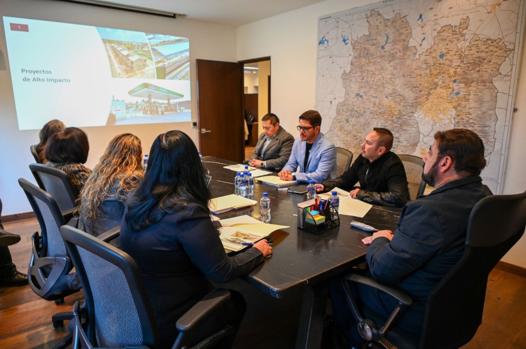 A business meeting taking place in a conference room, with seven professionals seated at a table, discussing projects. A presentation slide titled 'Proyectos de Alto Impacto' is displayed on a screen in the background.