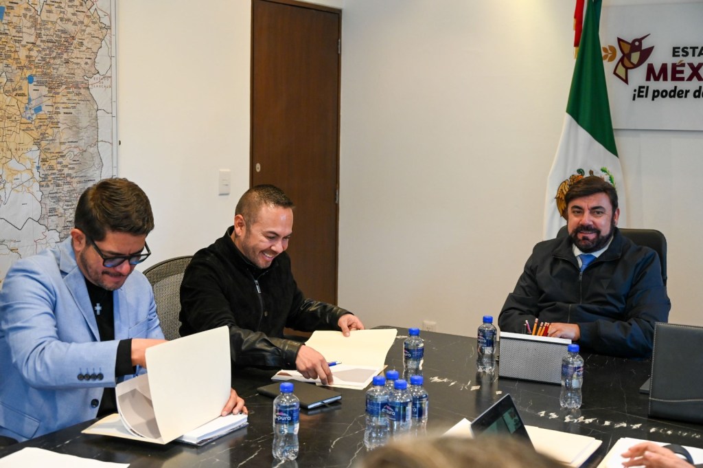 A group of three men sitting around a conference table with papers and water bottles, smiling and engaging in discussion. A map is visible on the wall behind them, along with the Mexican flag and a sign indicating the state of Mexico.