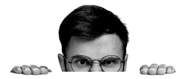 Close-up of a man with glasses peering over a surface, displaying a curious and slightly skeptical expression.