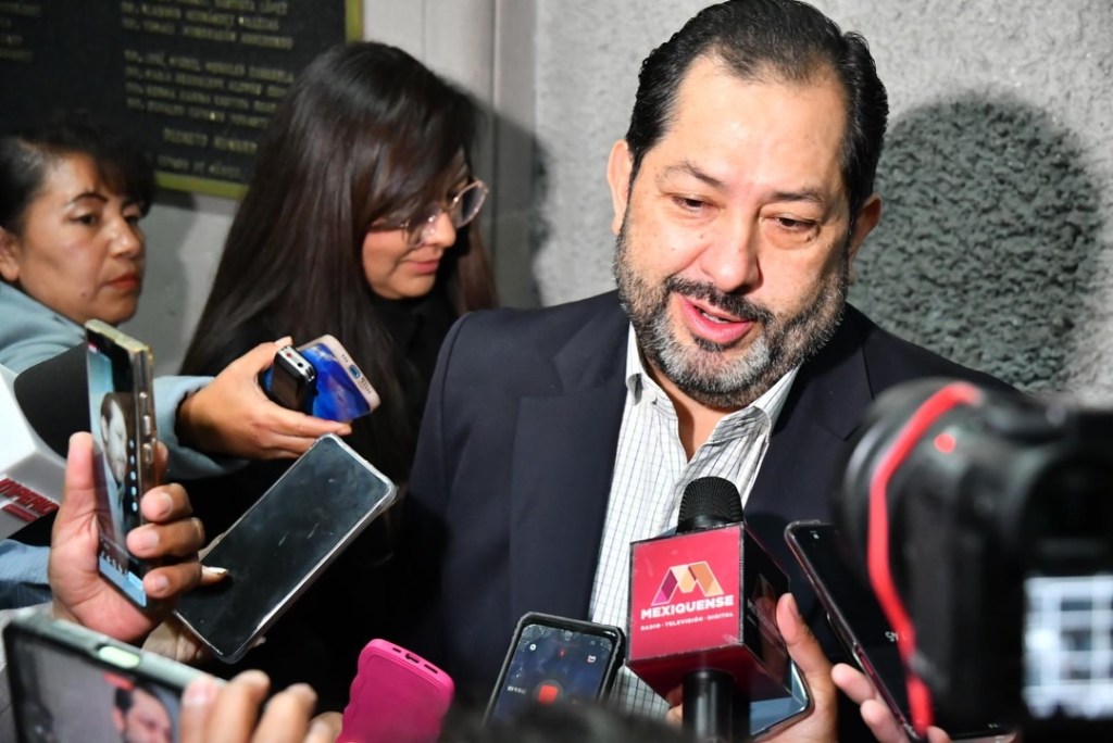 Un hombre con barba y saco de traje habla a un grupo de periodistas rodeado de cámaras y teléfonos móviles.