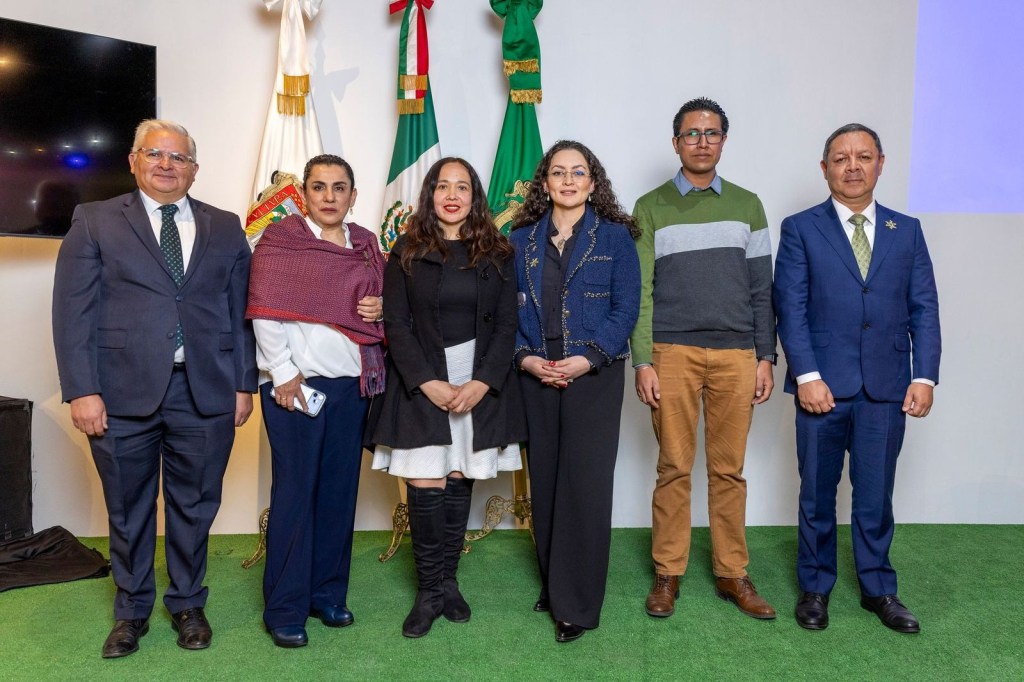 A group of six individuals, standing together in a formal setting with national flags in the background. They are dressed in a mix of formal and business casual attire.