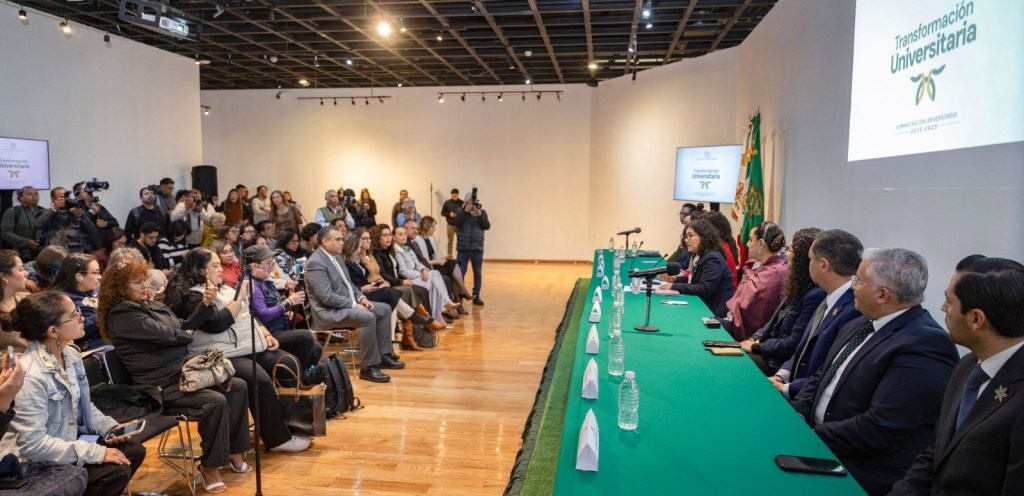 A conference or event in a spacious hall with a panel of speakers sitting behind a green table, addressing a large audience. Many attendees are seated and some are recording or taking pictures. A presentation screen is visible in the background, along with two flags.