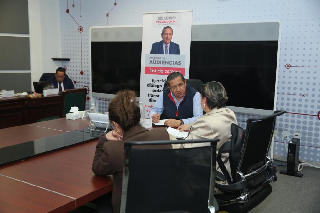 A meeting in a conference room with four individuals discussing over a table, with a banner behind one participant promoting a judicial program.