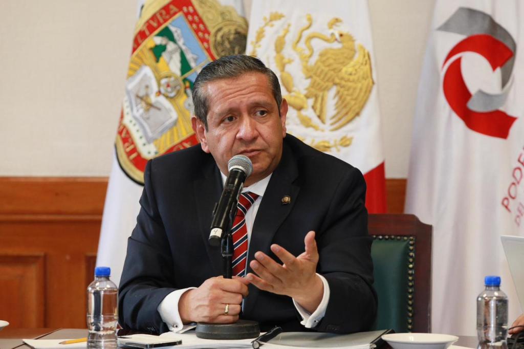 A man in a suit speaking at a table, microphone in hand, with flags in the background.