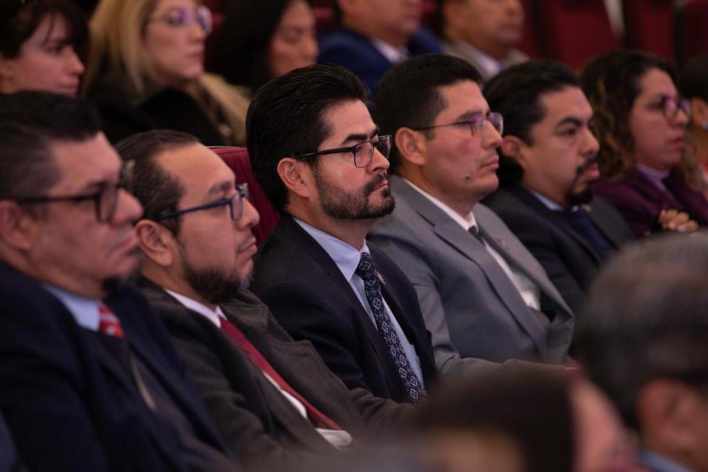 A group of people sitting in a conference or event setting, focused on the speaker. Several men in suits and ties are visible, exhibiting a serious demeanor.