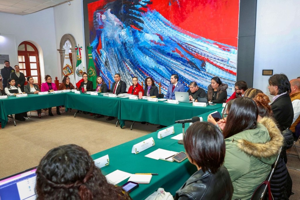 A meeting in a conference room with a large mural in the background, featuring multiple participants seated around a green table. The room includes flags on display, and attendees are engaged in discussion, with some using laptops and smartphones.
