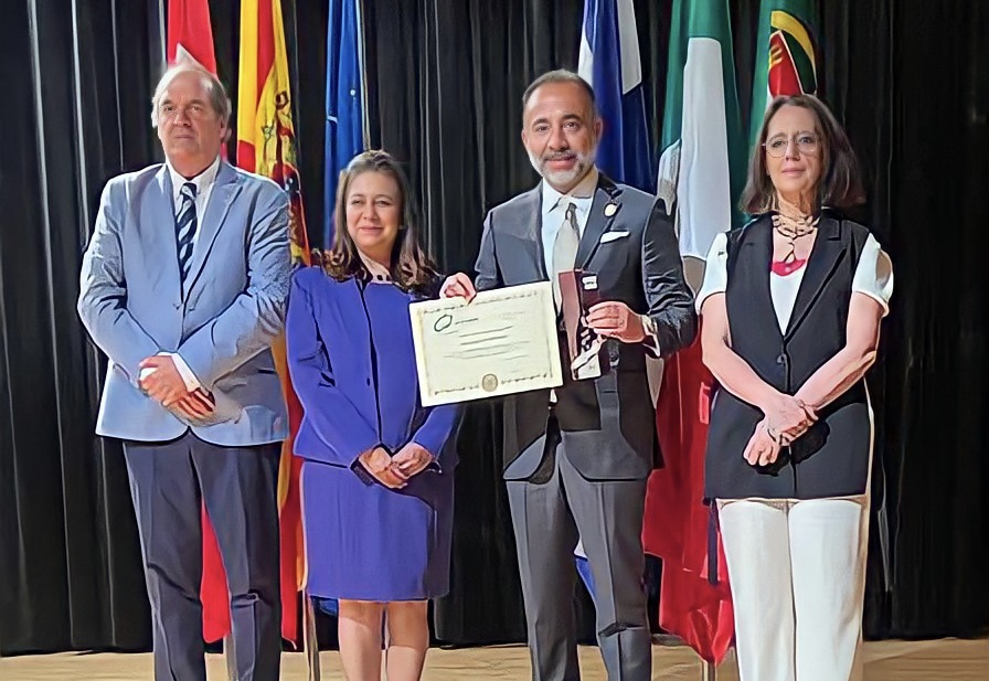Group of four people standing together on stage, holding a certificate. They are positioned in front of flags, showcasing a formal event.