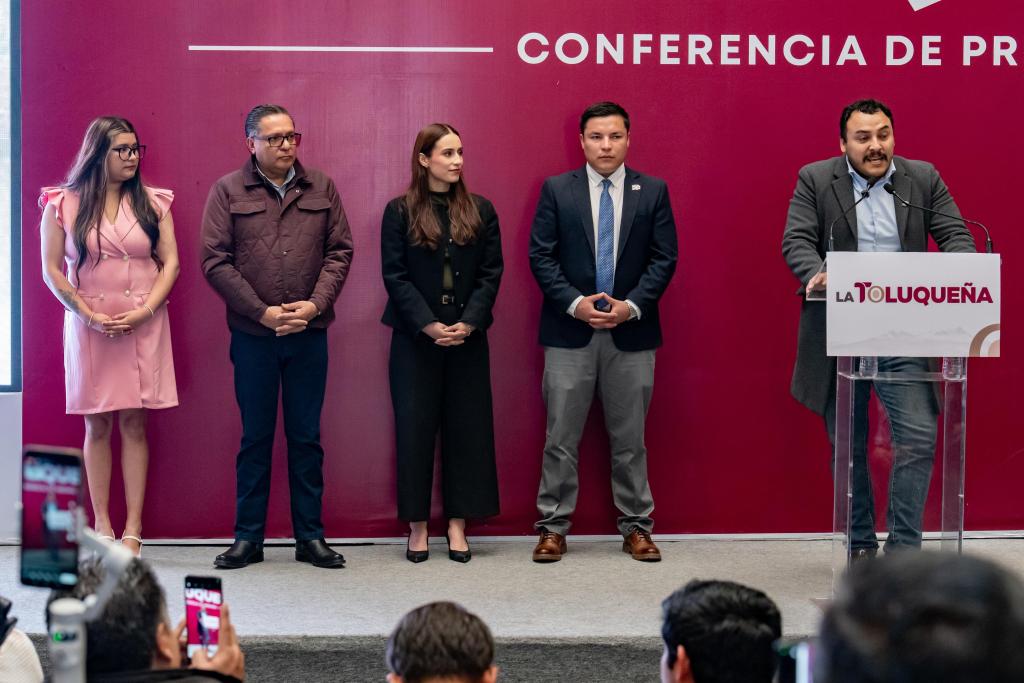 A press conference featuring five speakers, with one person speaking at a podium labeled 'LA TOLUQUEÑA' and a large banner behind them that reads 'CONFERENCIA DE PRENSA'. The group includes individuals in business attire and a woman in a pink dress.
