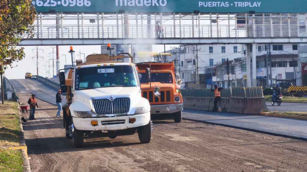 Two trucks on a construction site, one white and one orange, with workers in safety vests nearby. A pedestrian overpass and a sign are visible in the background.