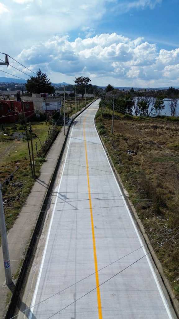 A long, winding road lined with electricity poles, bordered by grass and vegetation, under a partly cloudy sky.