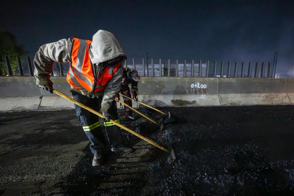 Trabajadores de la construcción trabajando en la rehabilitación de la carretera durante la noche, usando palas y llevando ropa de seguridad.