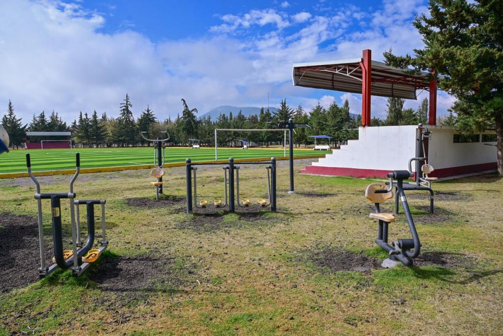 Outdoor fitness area with various exercise equipment on grassy terrain, under a blue sky with scattered clouds. A small grandstand can be seen in the background, near a green soccer field.