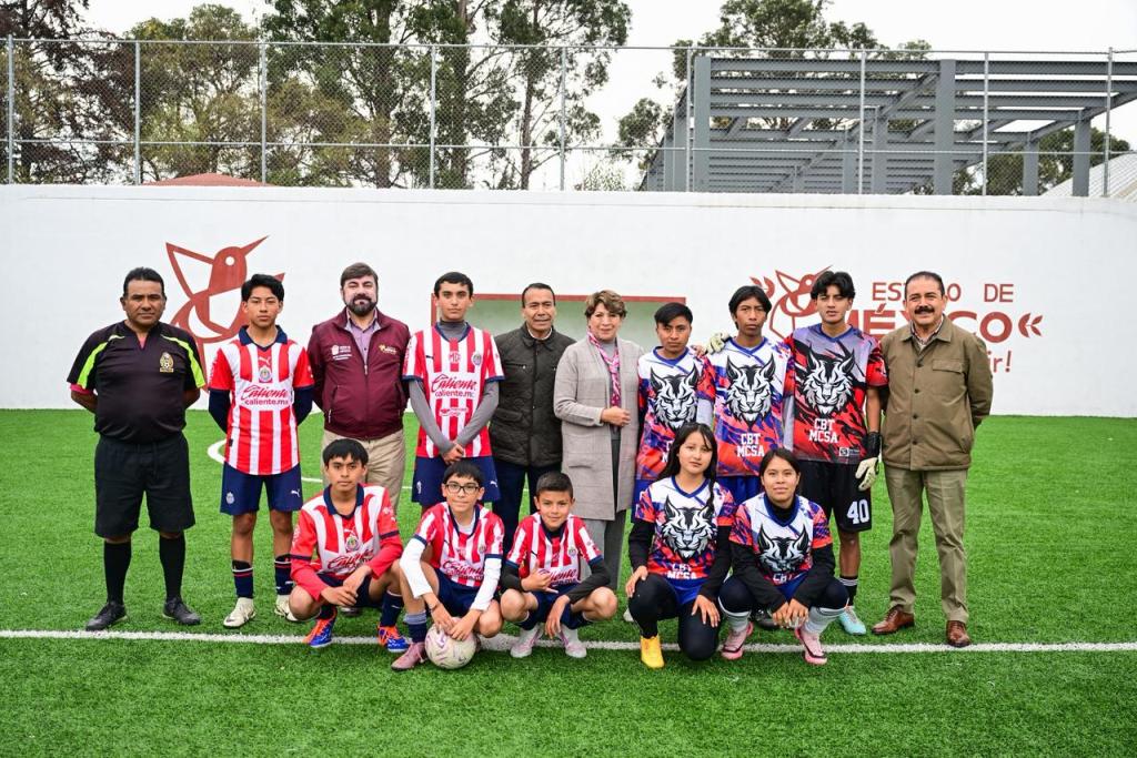 Group photo of a youth soccer team in red and white striped uniforms, with several adults in the background, standing on a green field.