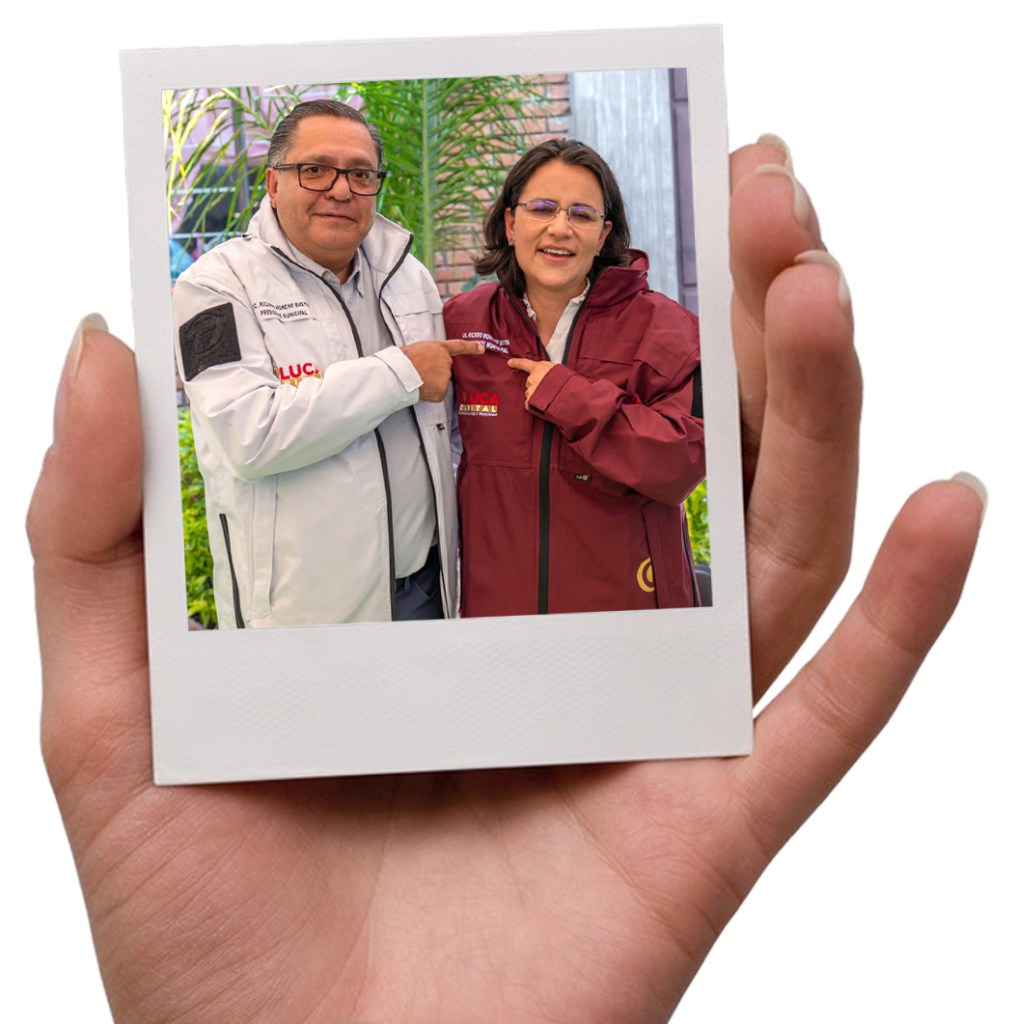 Una persona sosteniendo una fotografía de dos individuos sonriendo y posando juntos, uno vestido con chaqueta blanca y otro con chaqueta roja, en un entorno con plantas de fondo.