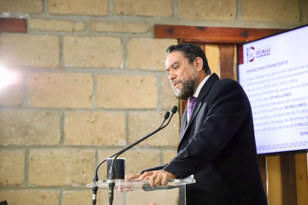 A man with a beard speaking at a podium, with a presentation displayed on a screen behind him. The background features a rustic stone wall.