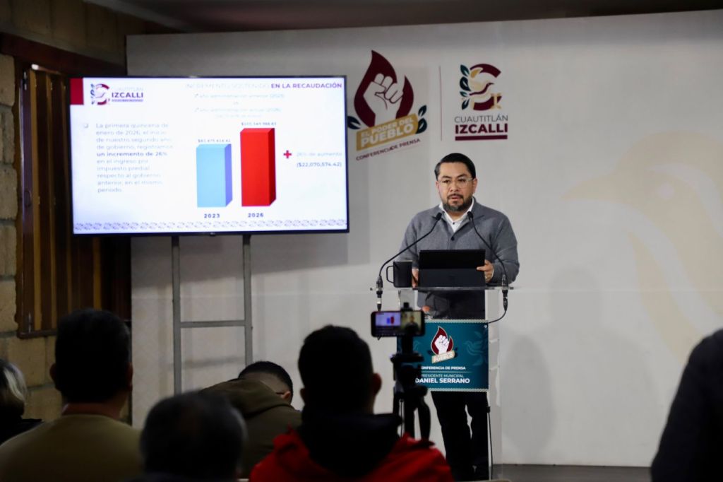 A man stands at a podium speaking during a press conference, with a presentation displayed on a screen highlighting financial data for the years 2023 and 2026. The backdrop features logos related to Cuautitlán Izcalli.
