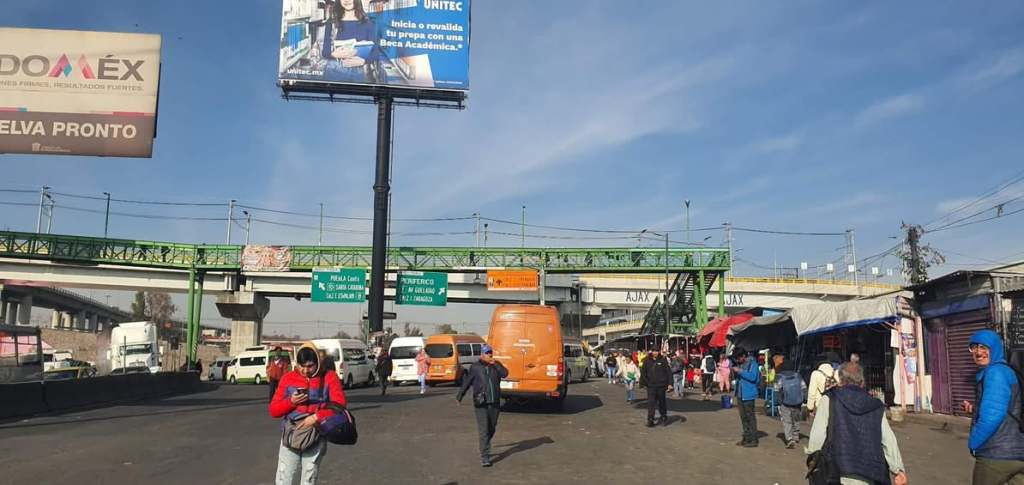 Busy urban street scene with people walking, vehicles, and billboards, under a clear blue sky.