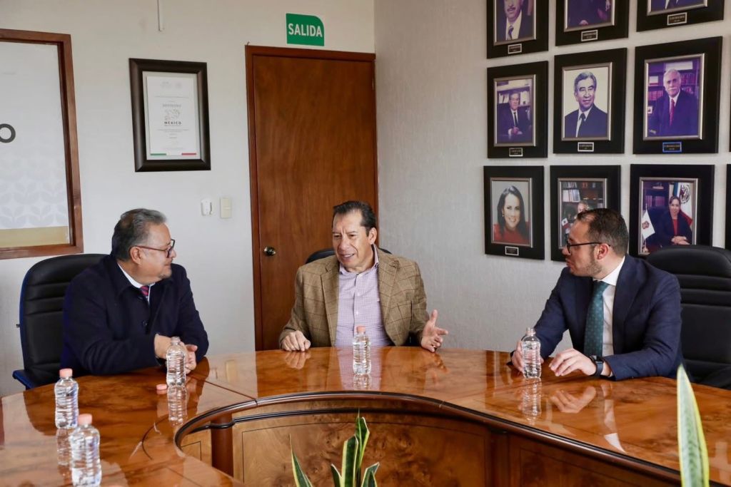 Three men engaged in conversation around a wooden table in a formal office setting, with framed photographs on the wall behind them.