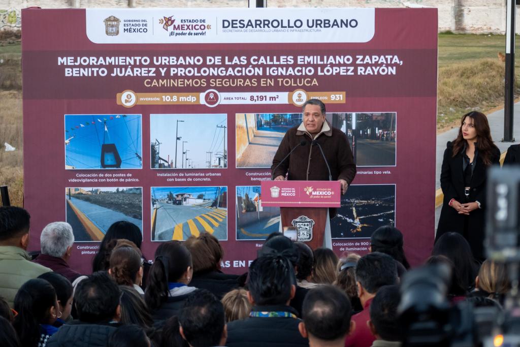 A public official speaks at a podium during an urban development event in Toluca, Mexico, with a large informational sign outlining improvements to local streets, including installations of streetlights and safety features, while an audience listens attentively.