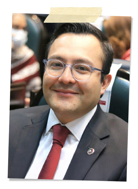 Retrato de un hombre sonriendo, con gafas, traje negro y corbata roja, sentado en un evento, con un fondo difuso de otros asistentes.