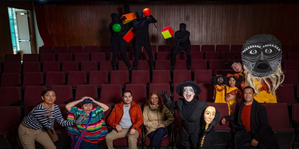 A diverse group of performers posing in a theater. In the front row, individuals in colorful costumes express excitement, while the back row features performers clad in black, holding props. The setting showcases layered seating and a wooden backdrop.