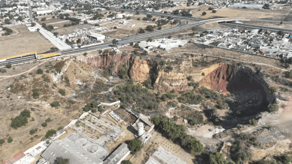 Aerial view of a large sinkhole surrounded by dry land, with buildings and roads in the background.