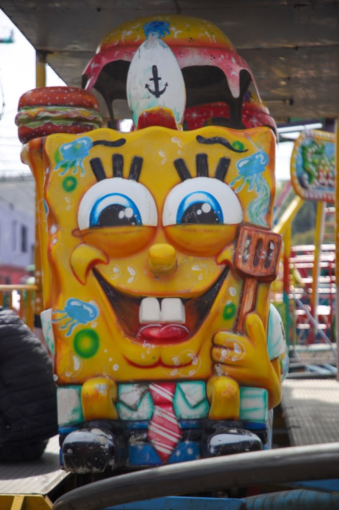 Colorful cartoon character resembling a sponge, wearing a chef's hat, holding a spatula, surrounded by funfair attractions.
