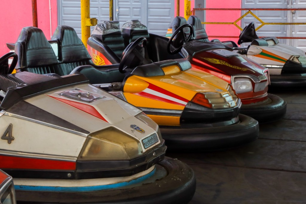 A row of colorful bumper cars parked on a wooden floor, featuring various designs and colors, in an indoor amusement setting.