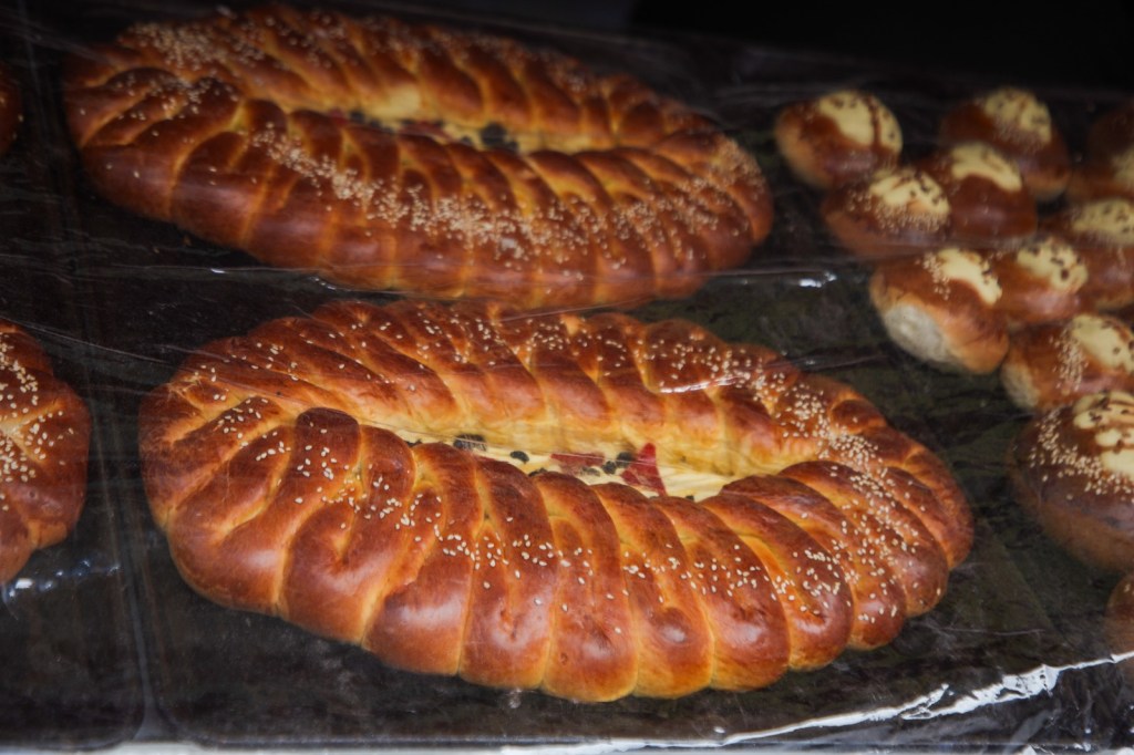 A display of freshly baked bread, featuring large, twisted loaves sprinkled with sesame seeds and smaller round buns glazed with creamy toppings.