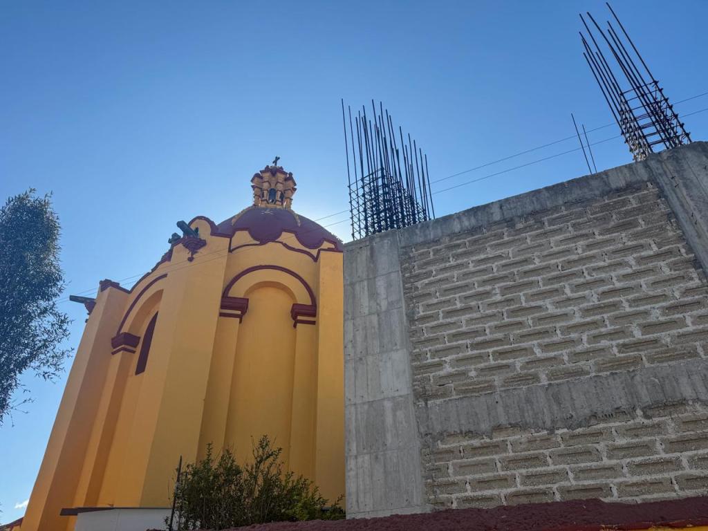 View of a yellow and red church with a dome, partially obscured by a concrete wall and construction rebar in the background against a clear blue sky.