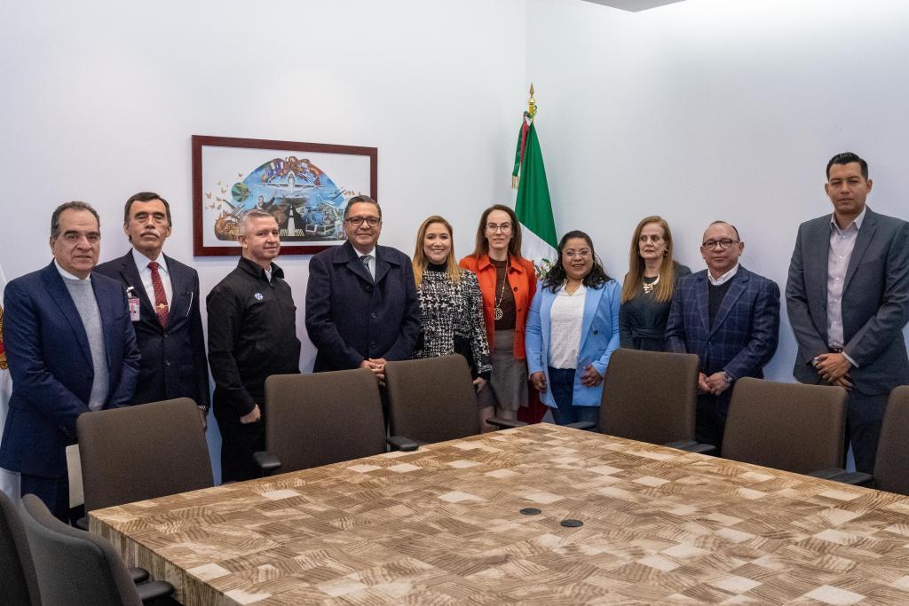 A group of twelve people standing in a meeting room, posing for a photo. A large wooden table is in the foreground, and a Mexican flag is visible in the background, along with a framed mural depicting cultural elements.