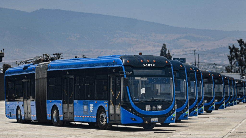 A row of blue electric buses parked in a yard with mountains in the background.