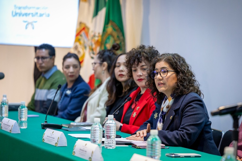 A panel of women speaking at a conference table, with a focus on the woman in a blue blazer and glasses. The background features flags and a presentation screen.