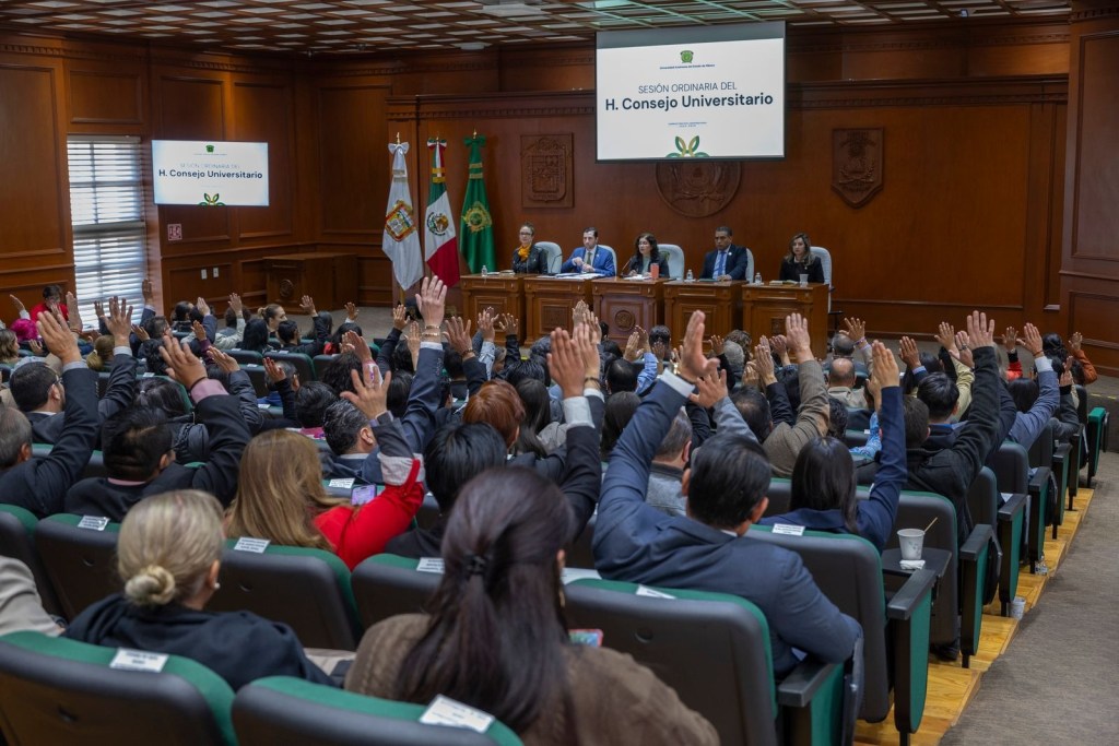 A formal meeting of the H. Consejo Universitario, featuring several attendees raising their hands to vote or indicate agreement, with flags and a panel of speakers in the background.