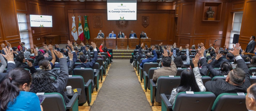 A university council meeting in a formal setting, with attendees raising their hands in a gesture of voting or agreement. The council is held in a wood-paneled room, featuring flags and a podium with speakers.