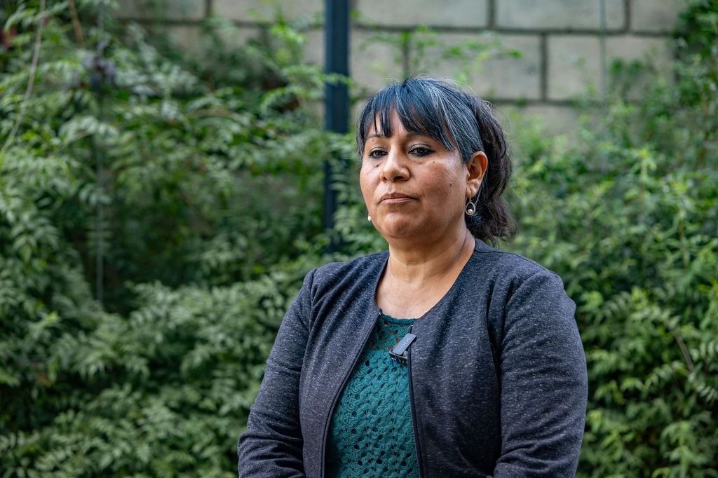 Una investigadora posando frente a un fondo de vegetación, vestida con una blusa verde y un abrigo oscuro, en un entorno natural.