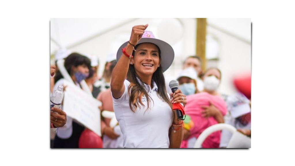 Mujer sonriendo y levantando el puño, hablando en un evento al aire libre, rodeada de un público que sostiene carteles y globos.