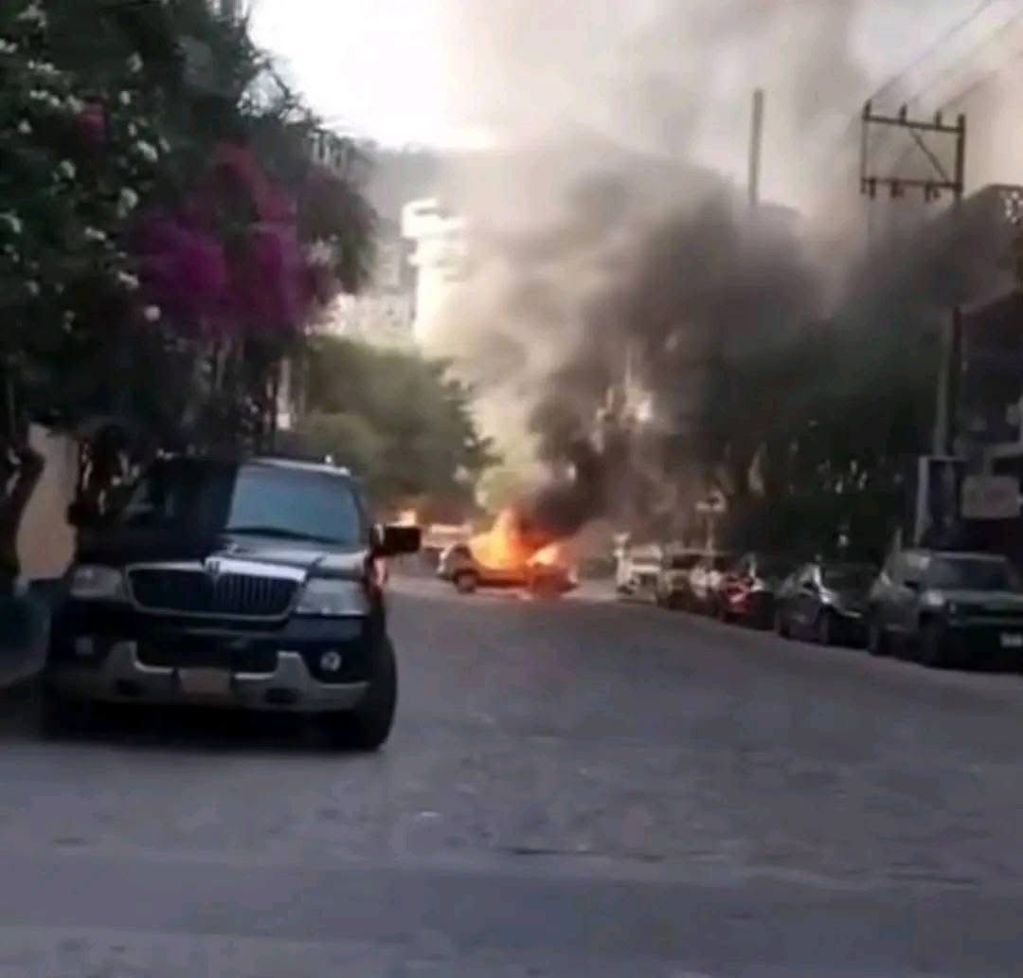 Un coche en llamas emitiendo humo en una calle con vehículos estacionados y vegetación alrededor.